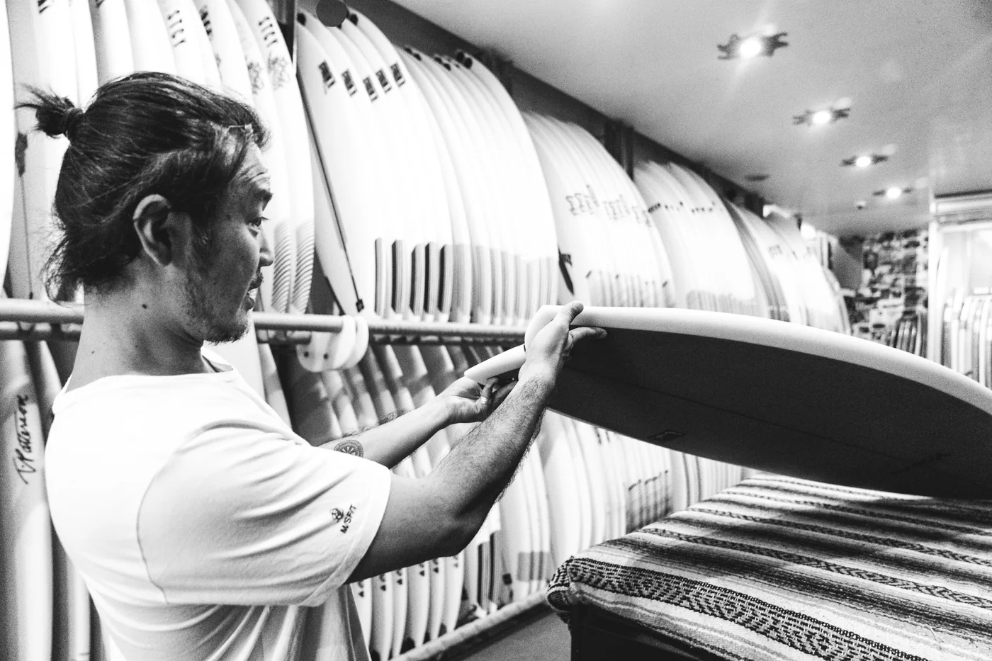 A person closely examining the rail of a surfboard in a room lined with boards